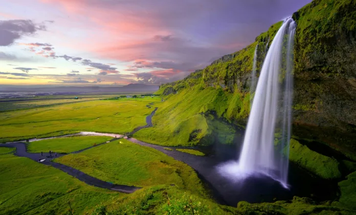 Scenic view of a tall waterfall cascading down a moss-covered cliff into a serene pool at sunset, creatively representing “waterfalls in Dallas” with vibrant green landscape and colorful sky.